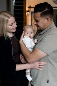 Baby zit tussen papa en mama in en krijgt een knuffel van beide ouders tegelijk.