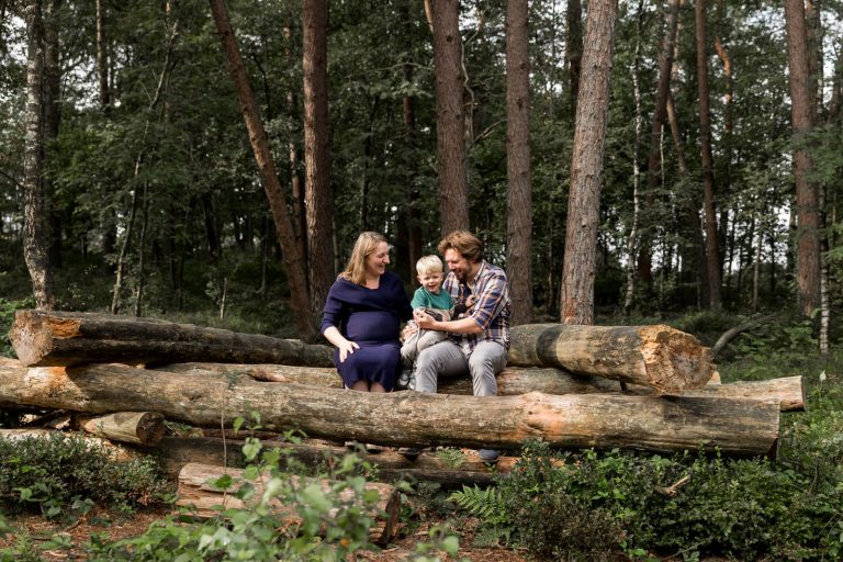 Gezin tijdens een zwangerschapsreportage op Het Leesten in Ugchelen, samen zittend op boomstammen en genietend van de natuur met hun zoon.