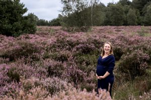 Mama met haar zwangere buik tussen de heide op Het Leesten in Ugchelen, met een brede glimlach terwijl ze haar buik vasthoudt en straalt van geluk.